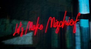 Glowing red neon light sign with cursive text 'Let's Make Mischief' on textured dark wall representing playful inappropriate humor